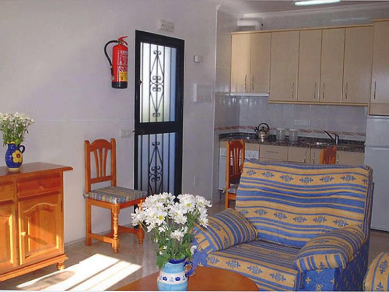 Kleines Wohnzimmer mit blau-gelbstreifigem Sofa, Holzmöbeln, weißen Wänden und offener Küche.