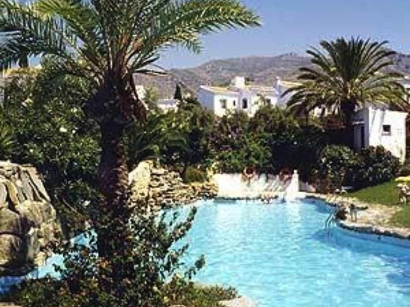 Klares Schwimmbecken mit Palme und Felsen, umgeben von bepflanztem Garten und weißen Gebäuden.