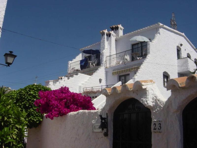 Weißes Haus mit Rundbögen, Balkonen und pinker Bougainvillea bei klarem Himmel.