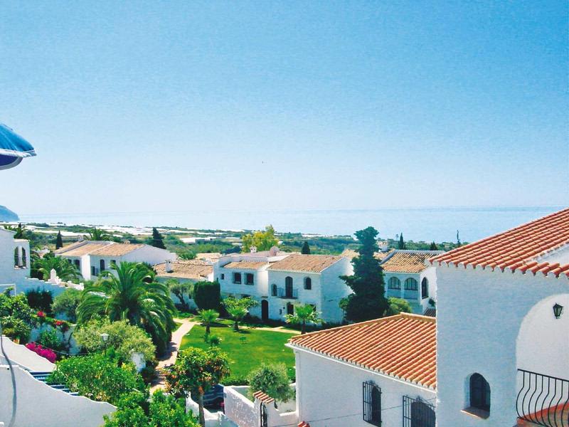 Helle Häuser mit roten Ziegeldächern, grüner Garten und blauer Himmel im Hintergrund.