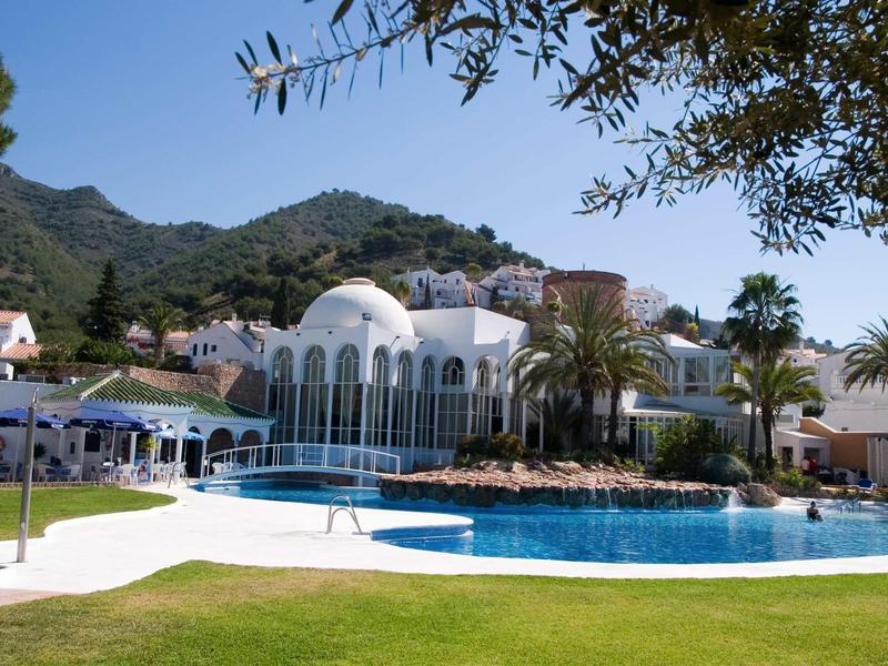 Weißes Hotel mit großen Bogenfenstern, Palmen und lebhaft blauem Pool vor Bergkulisse.