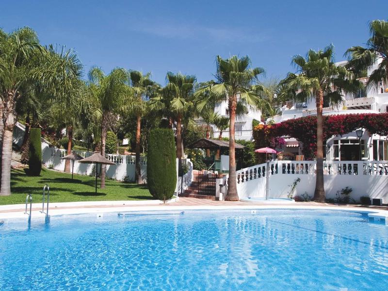 Blauer Swimmingpool mit weißen Geländern, umgeben von Palmen und Grünflächen unter blauem Himmel.