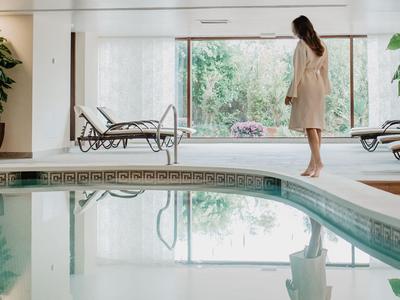 Frau steht am Rand eines Innenpools mit Liegestühlen und großen Fenstern im Hintergrund.