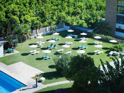 Grüner Garten mit weißen Sonnenschirmen, blauer Pool links, umgeben von Bäumen und Gebäuden.