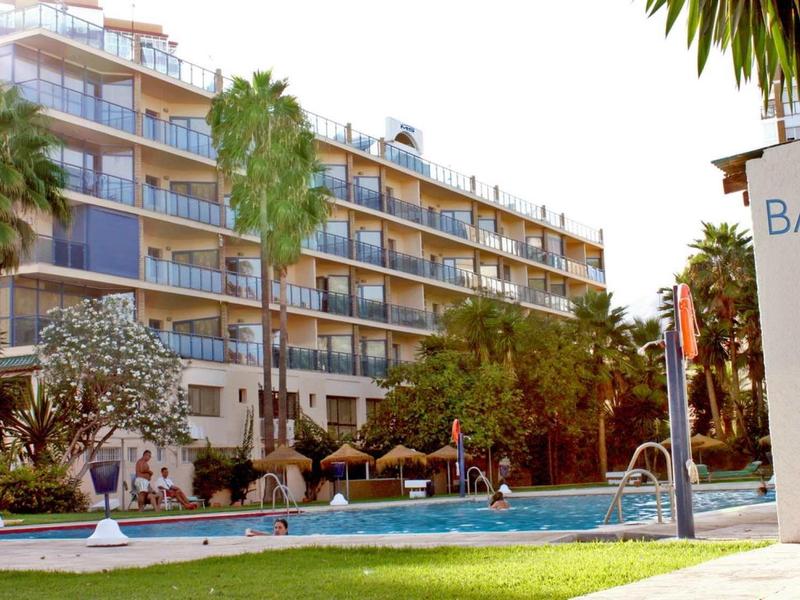 Mehrstöckiges Hotelgebäude mit Balkon, Garten, Pool und Palmen im sonnigen Klima.