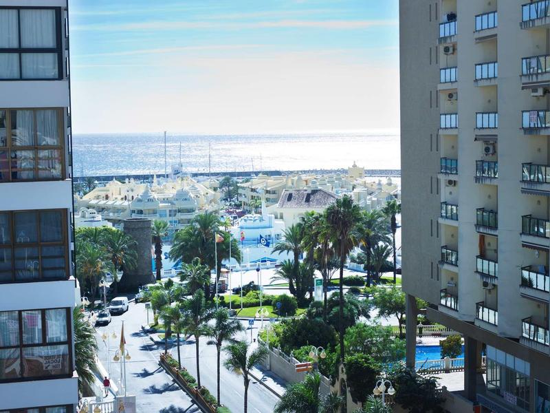 Blick auf Hafen mit Yachten und Palmen zwischen hohen Wohngebäuden bei klarem Himmel.