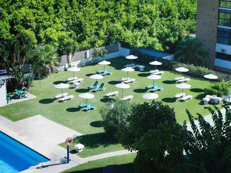 Grüner Garten mit weißen Sonnenschirmen, blauer Pool links, umgeben von Bäumen und Gebäuden.