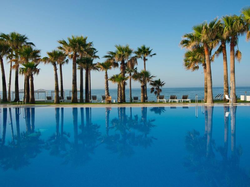 Großer, blauer Pool mit Palmen am Ufer und klarem Himmel im Hintergrund.