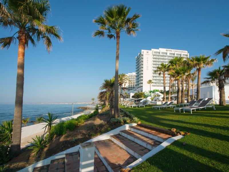 Strandpromenade mit Palmen, grünem Rasen und weißen Gebäuden unter klarem blauem Himmel.