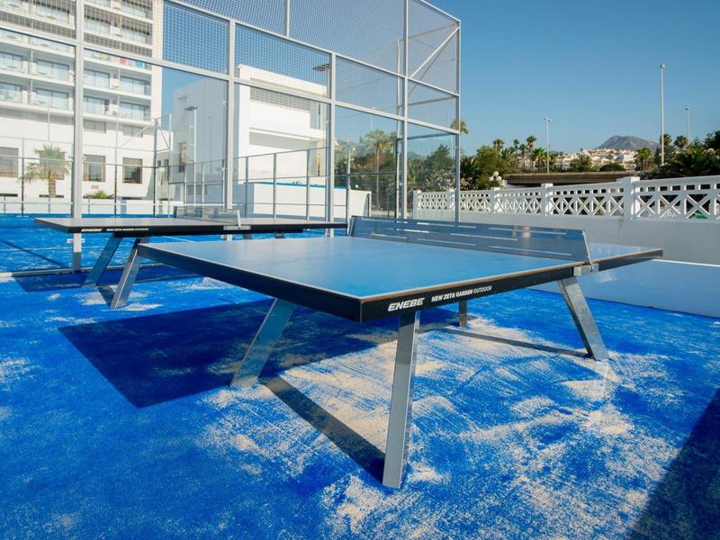 Blauer Tischtennistisch mit Metallbeinen auf blauem Boden im Freien bei sonnigem Wetter.