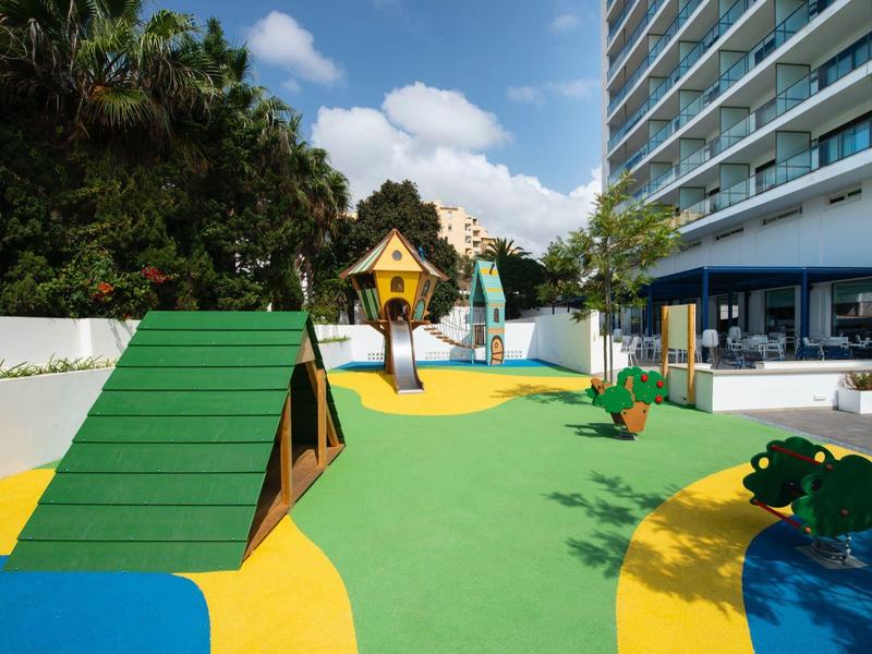 Bunter Kinderspielplatz mit Rutsche, Klettergerüst und grünem, gelbem Bodenbelag neben Gebäude.