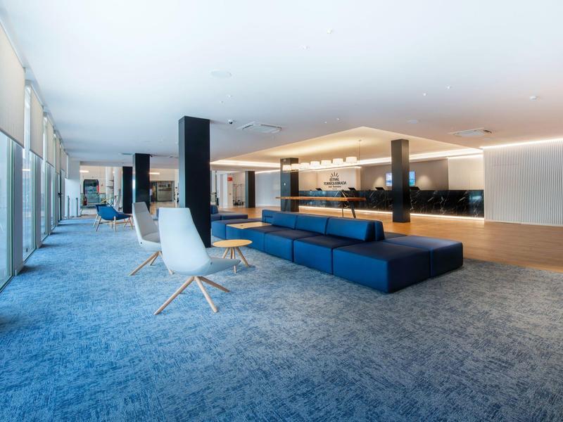 Moderner Wartebereich mit blauen Sitzbänken, weißen Stühlen und großen Fenstern an der Seite.