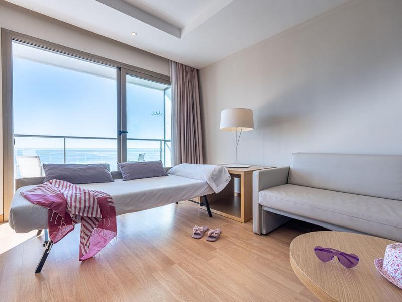 Helles Zimmer mit Liege, Sofa, seitlichem Tisch, Tischlampe, Balkon und Meerblick am Horizont.