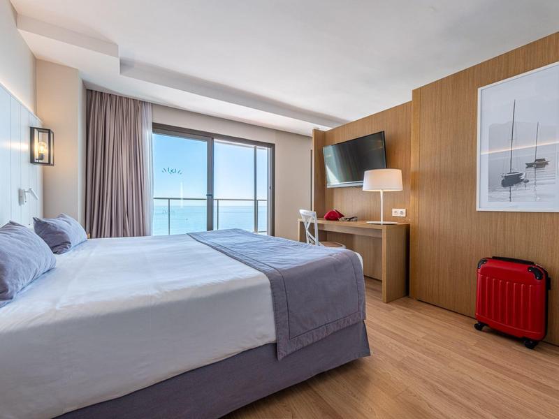 Hell eingerichtetes Hotelzimmer mit Doppelbett, Schreibtisch, Fenster mit Blick auf das Meer und rotem Koffer.