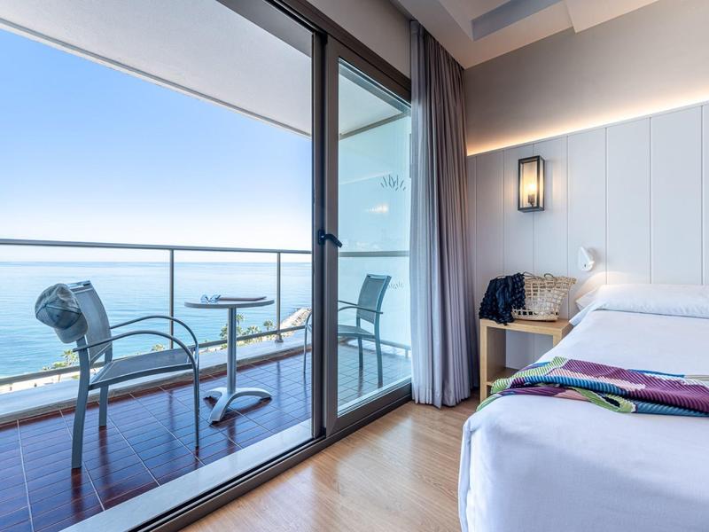 Modernes Hotelzimmer mit Doppelbett und großer Glasfront, Balkon mit Stühlen und Meerblick.