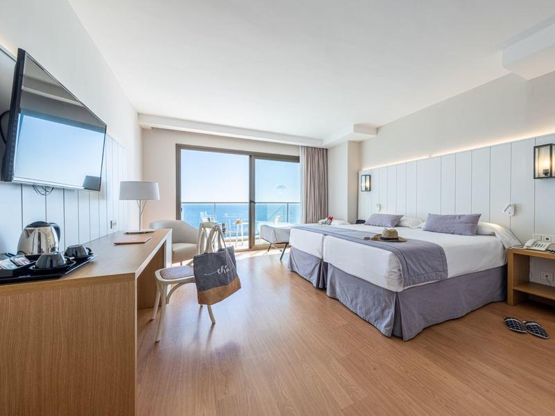 Helles Hotelzimmer mit Doppelbett, Holzboden, Schreibtisch, Fernseher und Balkon mit Meeresblick.