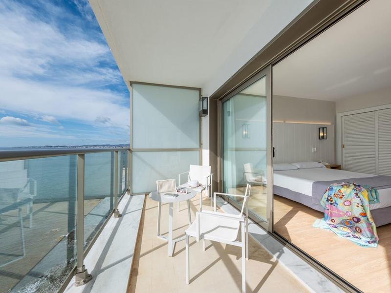 Balkon mit Glasgeländer, weißen Stühlen, angrenzendem Schlafzimmer, heller Himmel und Meerblick.