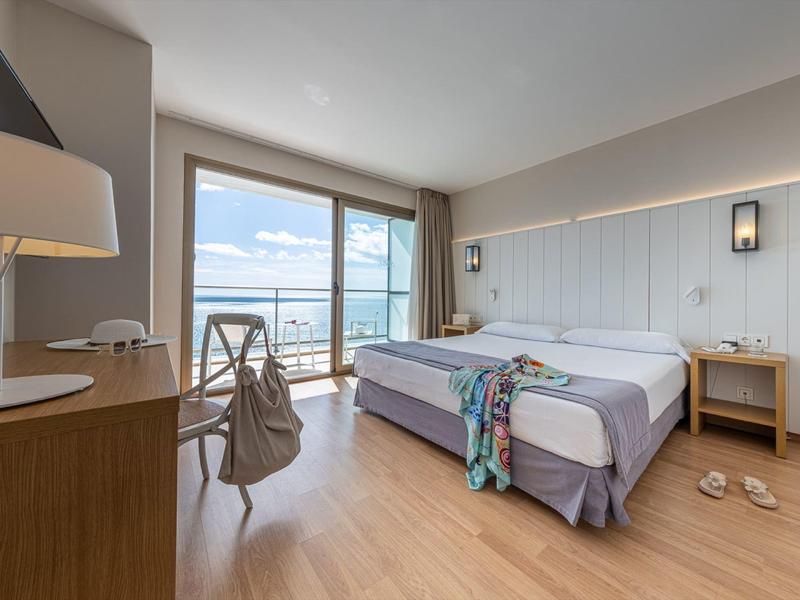 Helles Hotelzimmer mit Holzfußboden, Doppelbett, Balkon und Meerblick bei klarem Himmel.