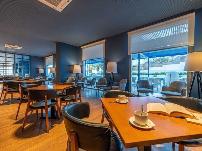 Modernes Café mit Holztischen, schwarzen Stühlen und großen Fenstern, viel Tageslicht.