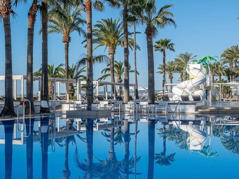 Blauer Pool mit Rutschen und Palmen, daneben weiße Liegestühle und Sonnenschirme.