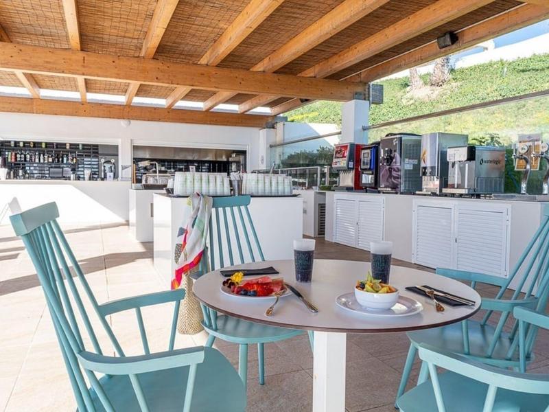 Hell belehrtes Außen-Café mit Holzdach, runden Tischen, türkisfarbigen Stühlen und Kaffeeautomaten.