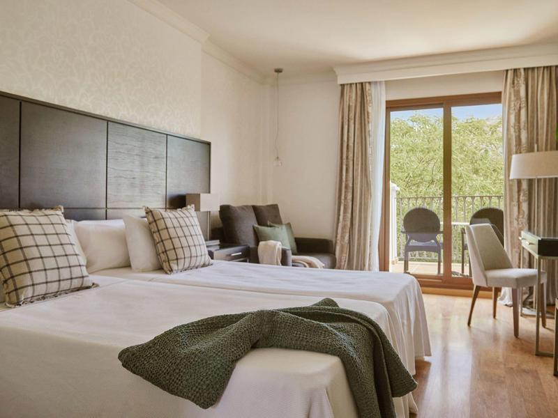 Hotelzimmer mit zwei Einzelbetten, Sesseln, Schreibtisch und Balkon mit Blick ins Grüne.