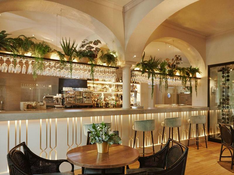 Innenraum einer stilvollen Bar mit geschwungenen Säulen, Pflanzen und Holzstühlen am runden Tisch.