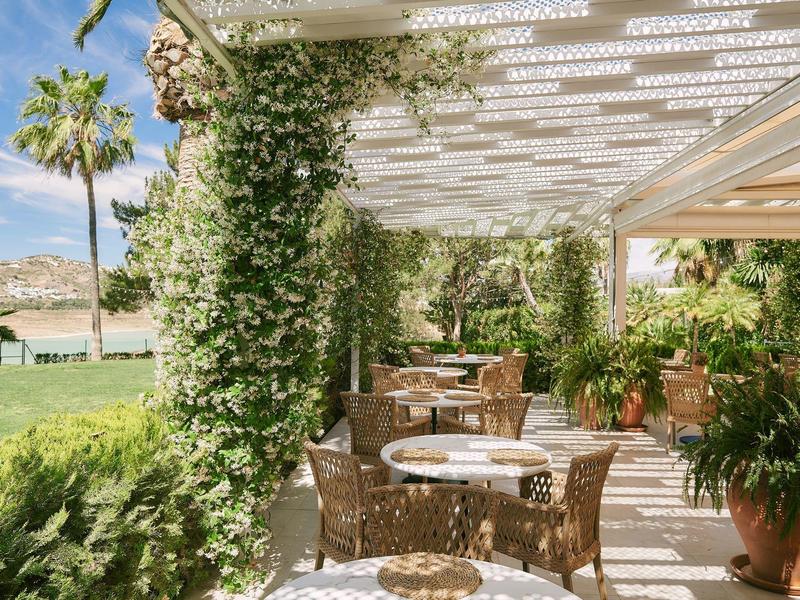 Terrasse mit runden Tischen, Stühlen und üppigem Grün an sonnigem Tag mit Blick auf See und Berge.