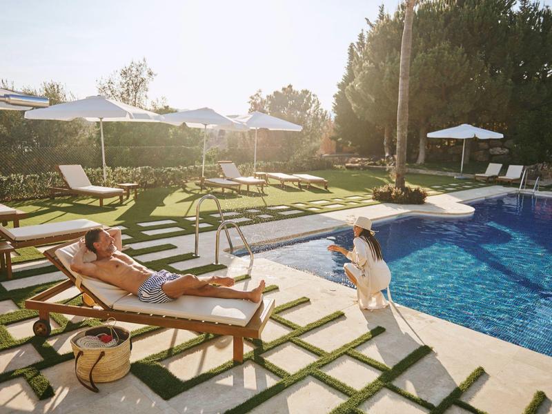 Sonnige Poollandschaft mit Liegestühlen, Sonnenschirmen, Pflanzen und zwei entspannenden Personen.