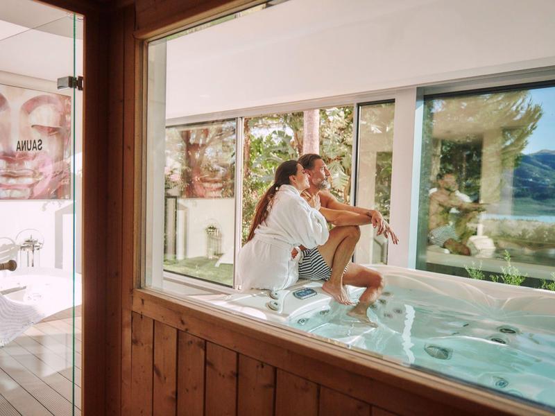 Frau entspannt im Whirlpool mit großer Glasfront und Bergblick in hellem, modernem Raum.