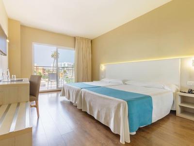 Helles Hotelzimmer mit Doppelbett, blauer Bettdecke, Schreibtisch, Fernseher und Balkon mit Blick.