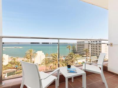 Balkon mit weißem Tisch und Stühlen, Blick auf Meer, Palmen und Gebäude bei klarem Himmel.