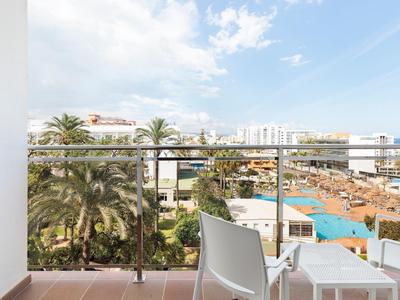 Balkon mit Stühlen, Blick auf Palmen, Pool und Gebäude unter blauem Himmel mit Wolken.