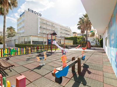 Spielplatz mit bunter Umzäunung, Rutsche, Wippe und Bänken vor einem weißen Gebäude mit Palmen.