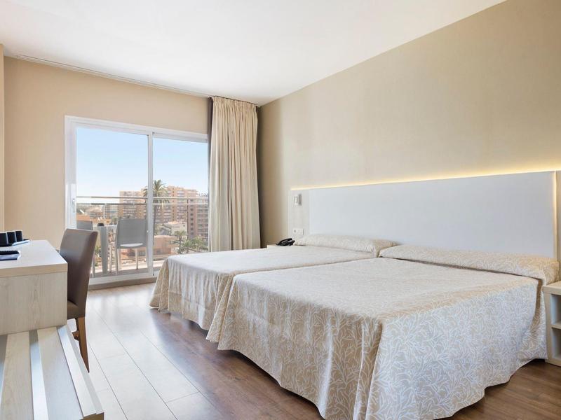 Hell eingerichtetes Hotelzimmer mit zwei Einzelbetten, Fenster mit Stadtblick und schlichter Möblierung.