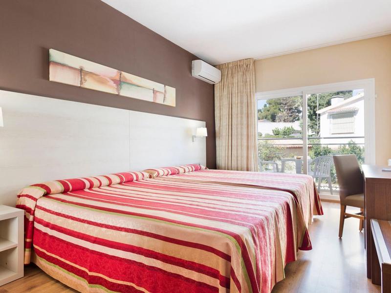 Helles Schlafzimmer mit Doppelbett, rot-gestreiften Bettdecken, brauner Wand und Balkon mit Gartenblick.