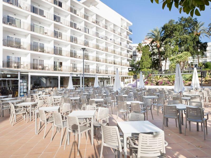 Große Terrasse mit vielen weißen Tischen und Stühlen vor einem weißem mehrstöckigen Hotel bei Tageslicht.