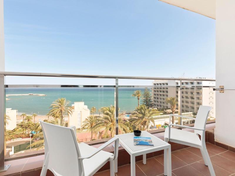 Balkon mit weißem Tisch und Stühlen, Blick auf Meer, Palmen und Gebäude bei klarem Himmel.