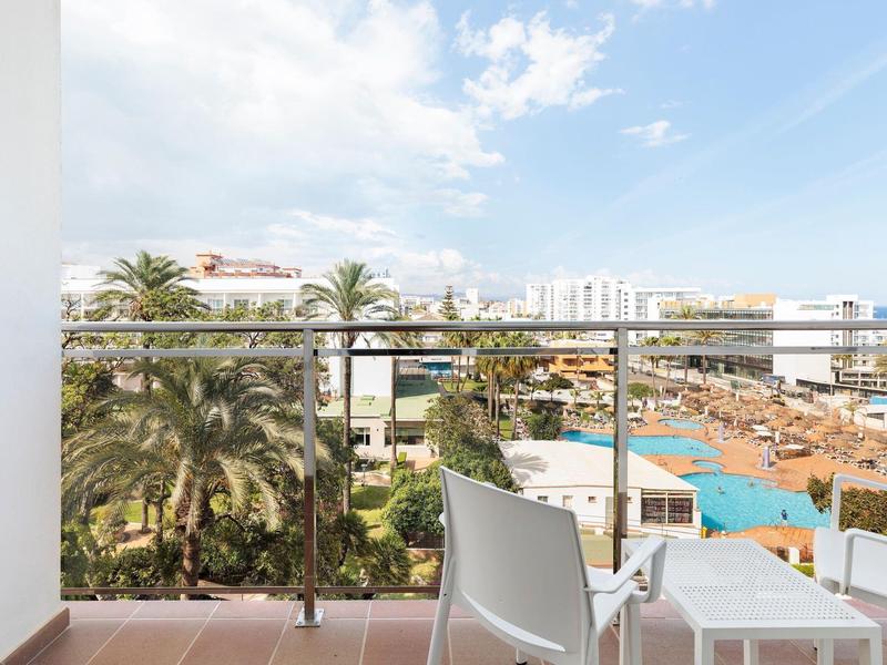 Balkon mit Stühlen, Blick auf Palmen, Pool und Gebäude unter blauem Himmel mit Wolken.