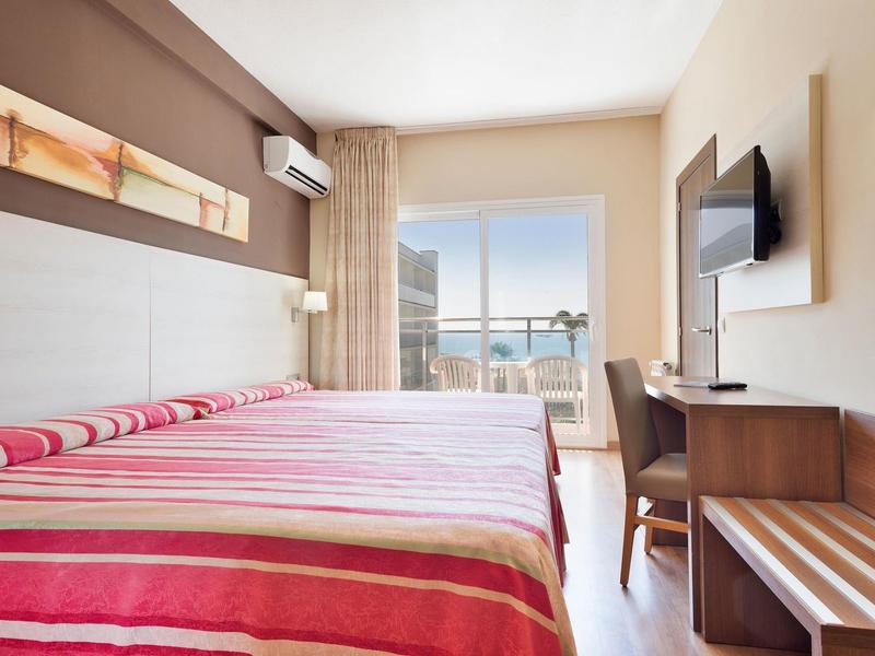 Helles Schlafzimmer mit Doppelbett, rot gestreifte Bettwäsche, Schreibtisch, Fernseher und Balkon mit Aussicht.