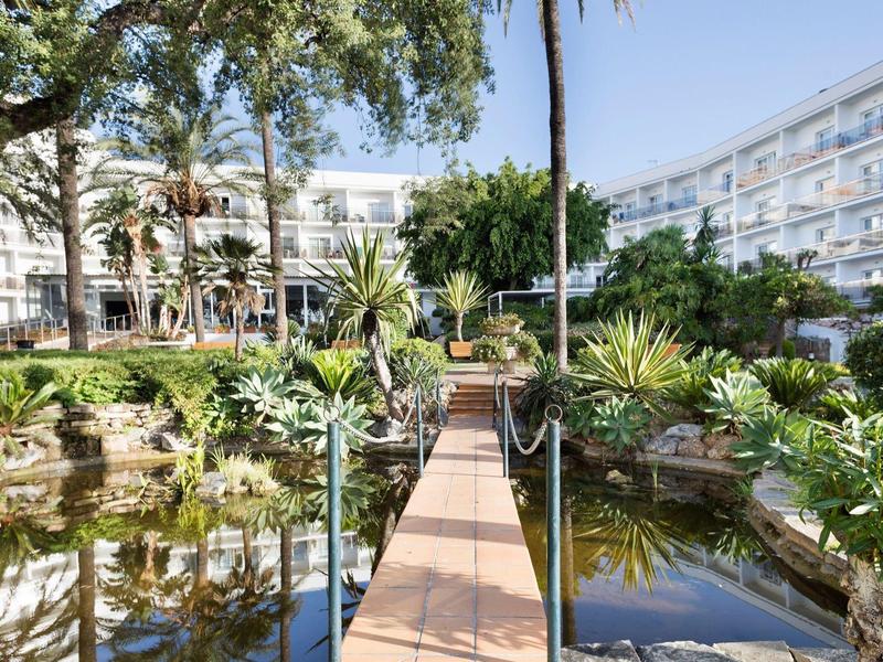 Holzbrücke führt über Teich zu weißem, mehrstöckigem Hotel mit grünen Palmen und Pflanzen.