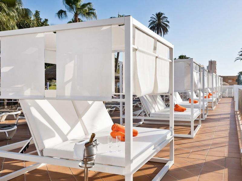 Reihen von weißen Lounge-Betten mit weißen Vorhängen und orangen Handtüchern bei Sonnenschein.