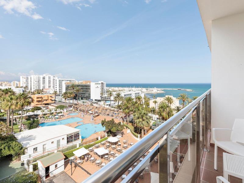 Blick vom Balkon auf Hotelanlage, Palmen, Strand und blaues Meer unter hellem Himmel.