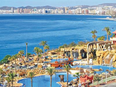 Küstenlandschaft mit blauem Meer, Palmen, Pool und Hafen in sonnigem, mediterranem Ambiente.