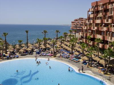 Großer runder Pool mit Schwimmern, Palmen, Liegen und mehrstöckigem Gebäude am Meer im Hintergrund.