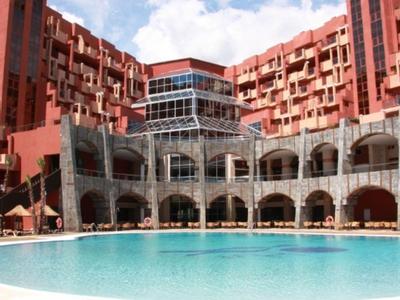 Gran piscina de hotel frente a un edificio rojo de varios pisos con ventanas arqueadas y terraza al aire libre.