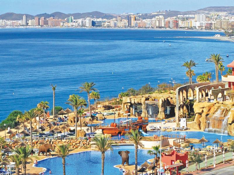 Küstenlandschaft mit blauem Meer, Palmen, Pool und Hafen in sonnigem, mediterranem Ambiente.