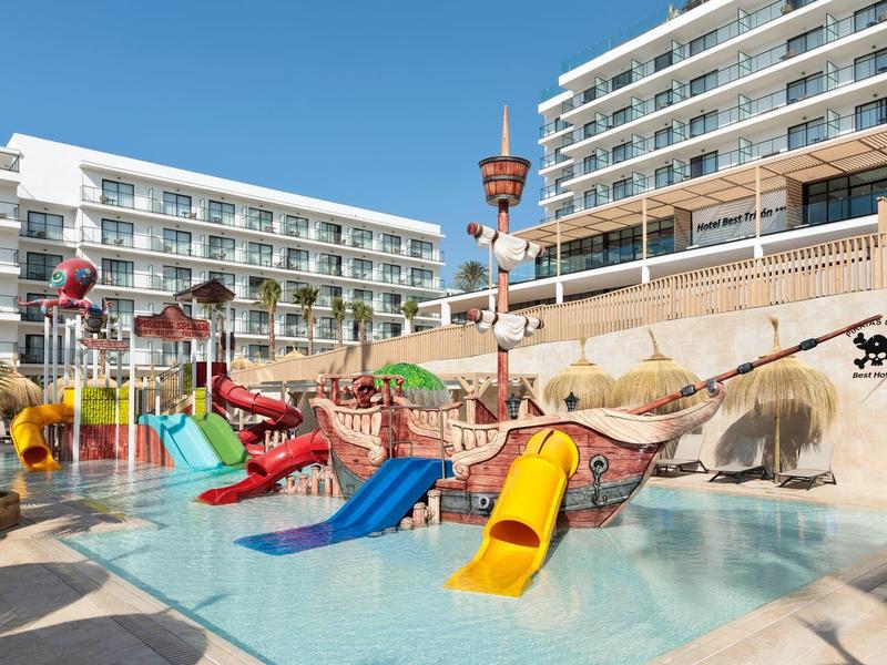 Kinderplanschbecken mit bunten Rutschen und Piratenschiff-Dekoration vor Hotels bei klarem Himmel.