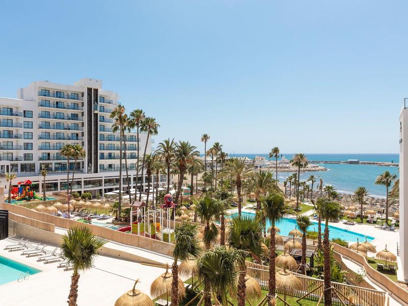 Hotelanlage mit Pools, Palmen und Meerblick unter klarem Himmel an sonnigem Tag.