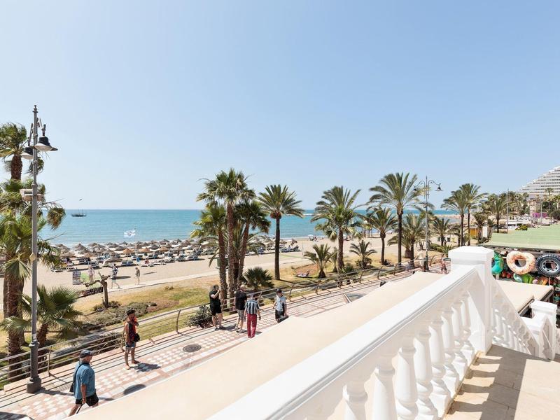 Strandpromenade mit Palmen, Fußgängern, Strand und Meer unter klarem blauen Himmel.